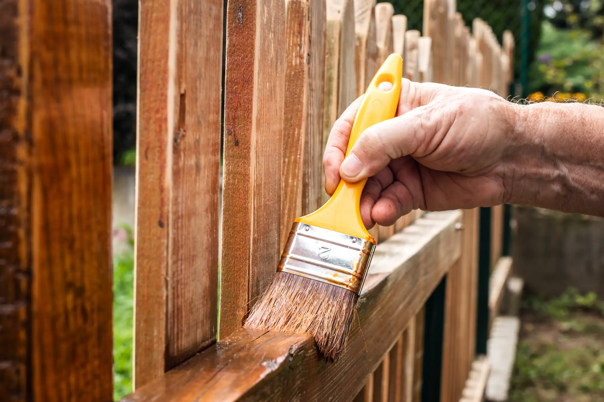 A hand with a yellow paintbrush applies wood stain to a fence by Fence Company Suffolk County, NY.