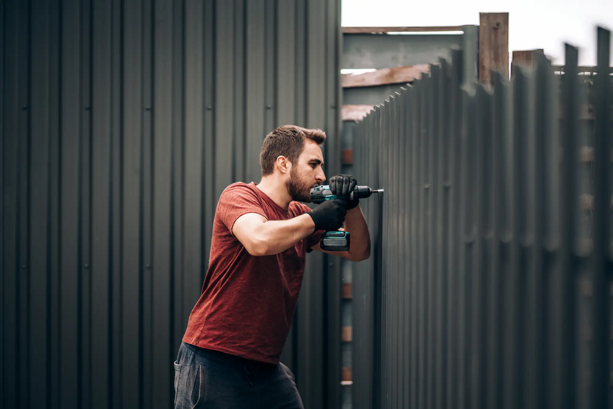 A man in a red shirt from a Fence Company Suffolk County uses a power drill to repair a metal fence.