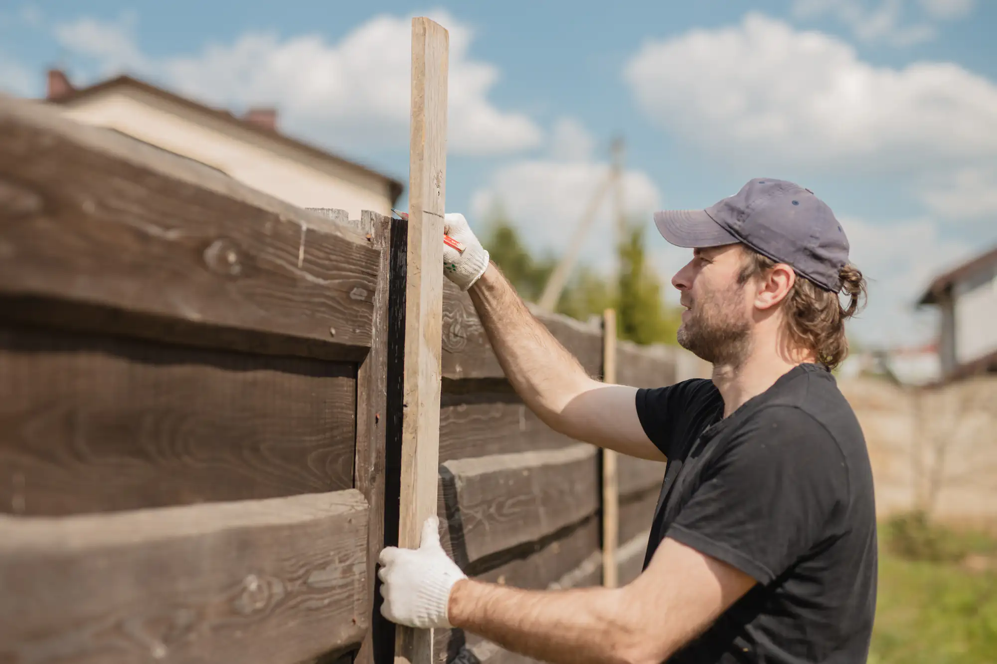 A man in a cap and gloves measures a wooden fence outdoors on a sunny day for Fence Company Suffolk County.