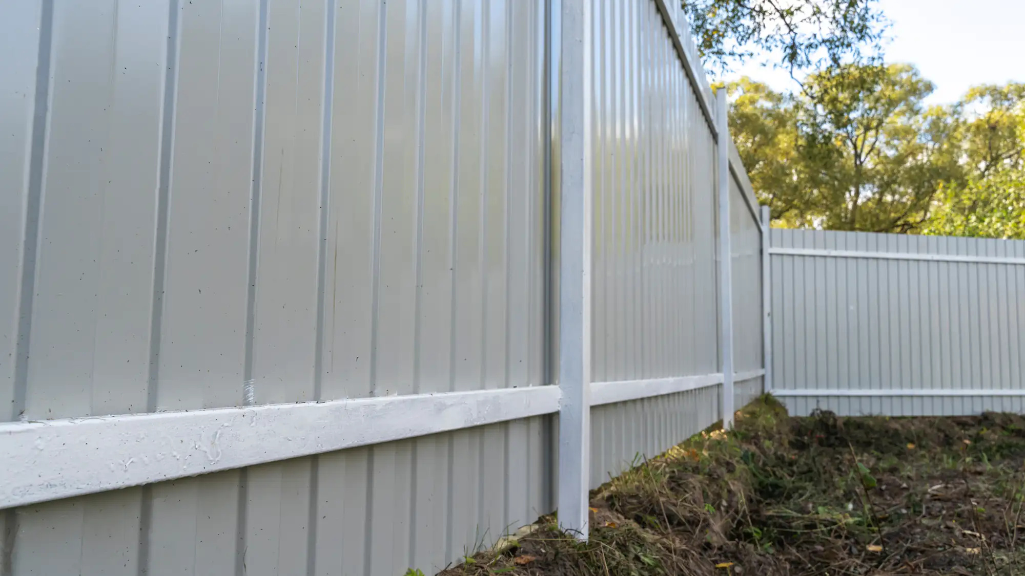 A close-up shows a tall, light gray metal fence by a top fence contractor Suffolk County with green trees behind.