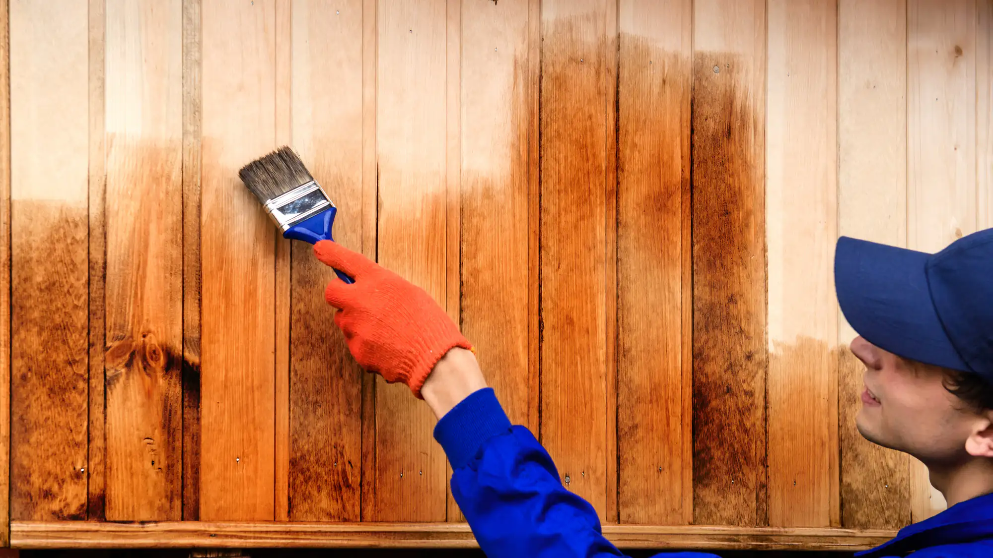 A person in blue and orange paints a wooden fence, showcasing skills like a Fence Company Suffolk County expert.