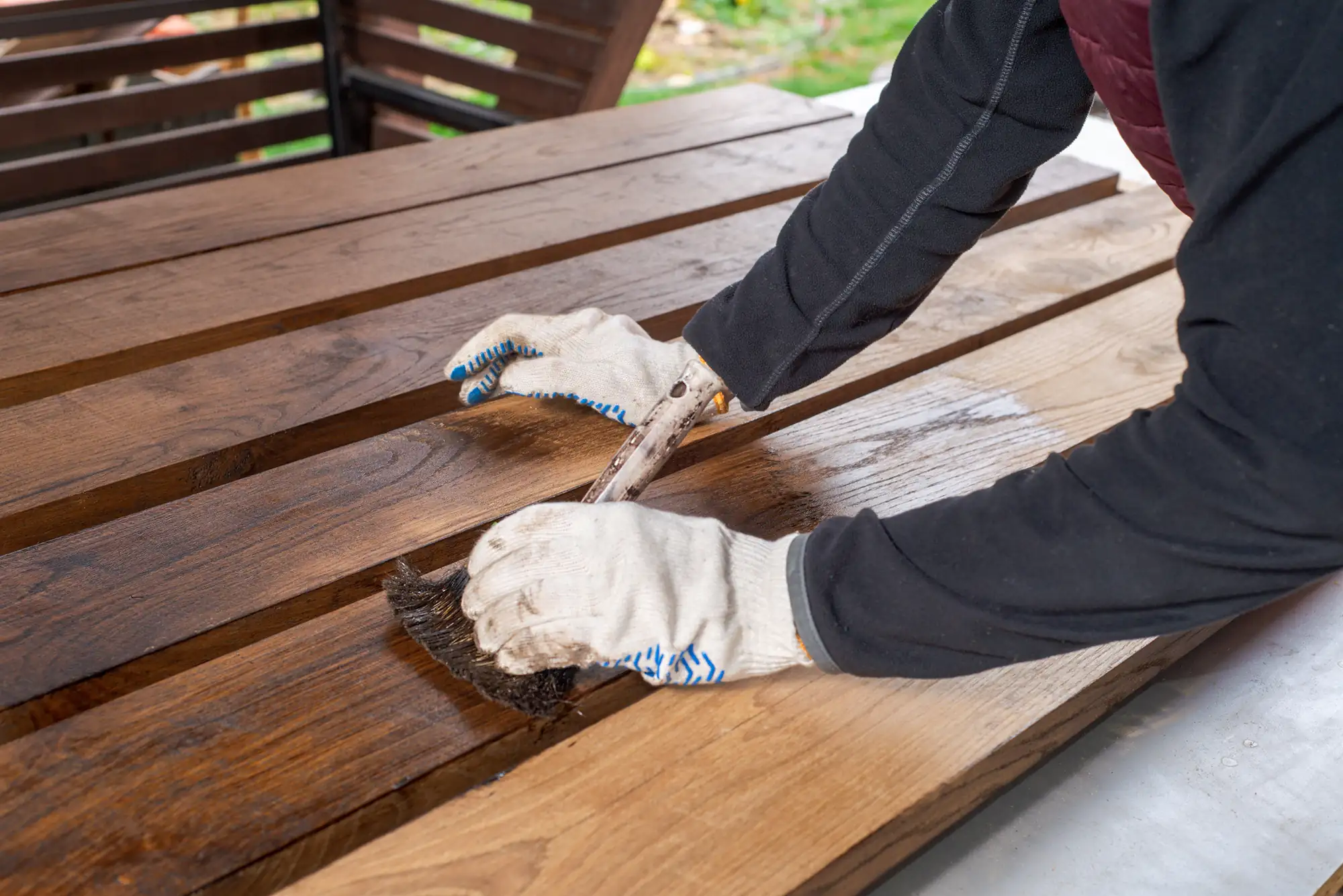 A person in gloves stains wooden planks outdoors, like a NY fence contractor Suffolk County at work.