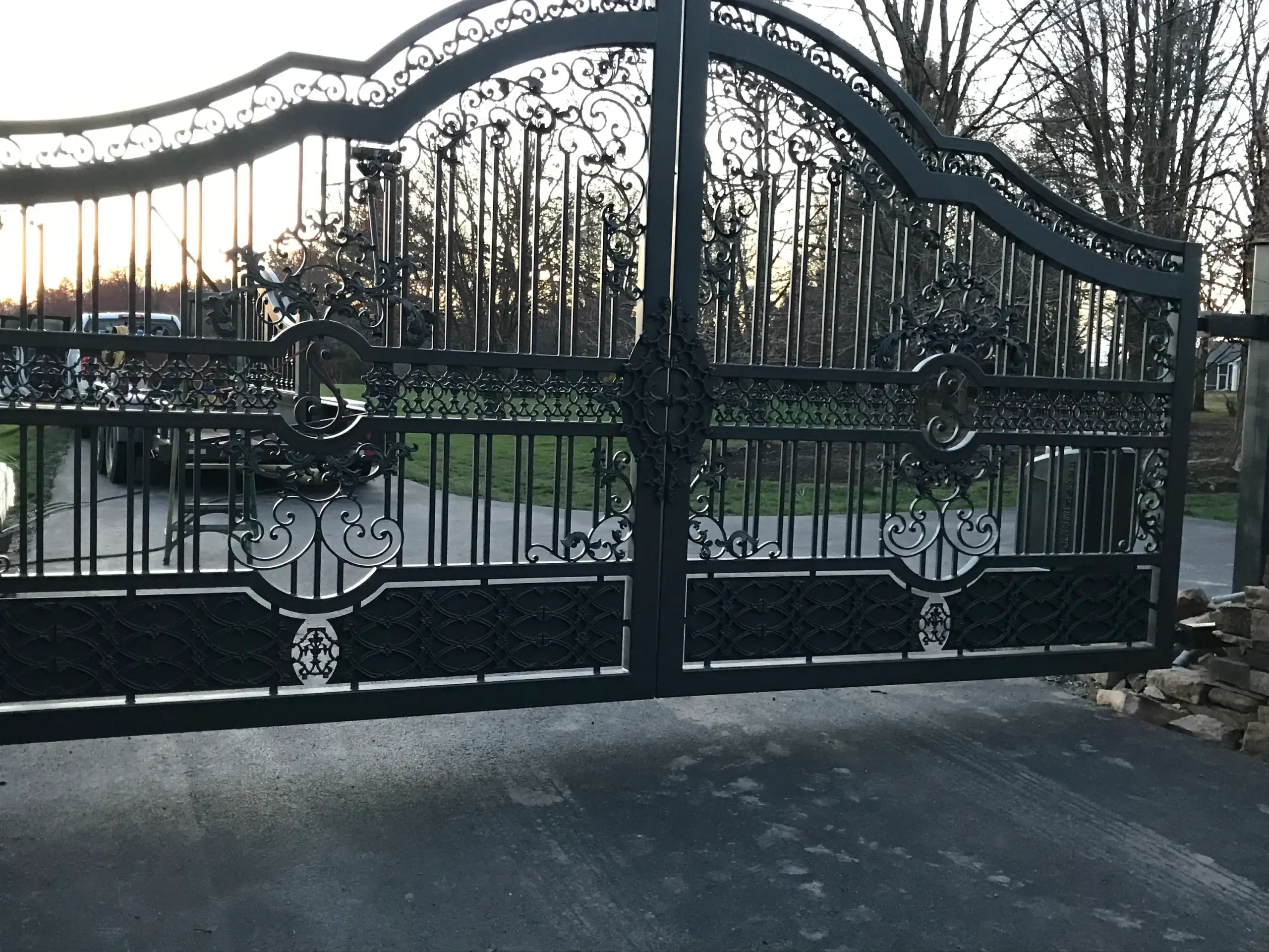 Ornate black wrought iron gate closed across a driveway, with green trees and grass in the background.